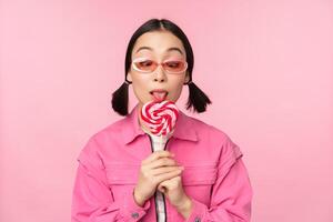 tonto y linda asiático hembra modelo paliza piruleta, comiendo caramelo dulce y sonriente, mirando entusiasmado, en pie terminado rosado antecedentes foto