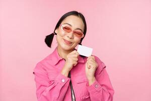 retrato de elegante, moderno asiático chica, muestra descuento, crédito tarjeta y Mira complacido, pago sin contacto, concepto de compras, en pie terminado rosado antecedentes foto