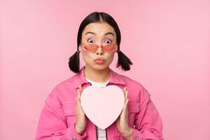 Cute asian girl showing heart shaped box with gift, looking surprised and excited, romantic present concept, wearing sunglasses, standing over pink background photo
