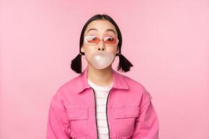 Stylish asian girl blowing bubblegum bubble, chewing gum, wearing sunglasses, posing against pink background photo