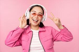 Dancing stylish asian girl listening music in headphones, posing against pink background photo