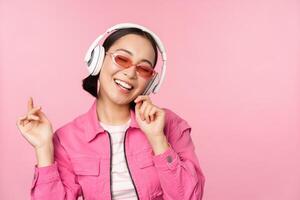 Dancing stylish asian girl listening music in headphones, posing against pink background photo