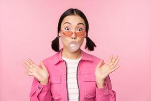 Stylish asian girl blowing bubblegum bubble, chewing gum, wearing sunglasses, posing against pink background photo