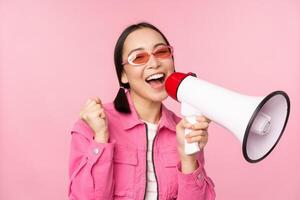 Attention, announcement concept. Enthusiastic asian girl shouting in megaphone, advertising with speaker, recruiting, standing over pink background photo