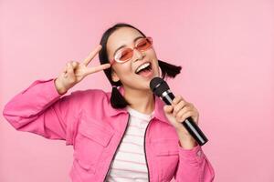 Happy beautiful asian girl singing with mic, using microphone, enjoying karaoke, posing against pink studio background photo