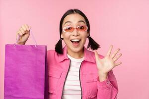 compras. elegante asiático niña en Gafas de sol, demostración bolso desde tienda y sonriente, recomendando rebaja promoción en almacenar, en pie terminado rosado antecedentes foto