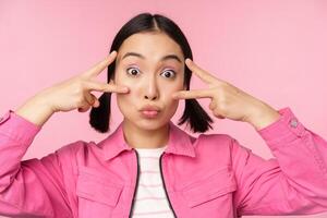 Close up of stylish asian girl smiles happy, shows peace v-sign, kawaii pose, posing against pink background photo