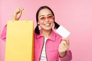 Happy young asian woman showing credit card for shopping, holding bag, buying on sale, going to the shop, store, standing over pink background photo