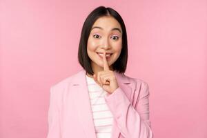 Hush, taboo concept. Portrait of asian businesswoman showing shush gesture, shhh sign, press finger to lips, standing over pink background in suit photo