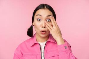 Close up portrait of young asian girl looking surprised, express amazement and wonder, peeking through fingers, standing over pink background photo