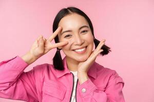 Close up of stylish asian girl smiles happy, shows peace v-sign, kawaii pose, posing against pink background photo