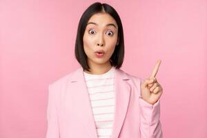 Got an idea. Young asian saleswoman, office manager raising finger, suggesting, wearing suit, posing against pink studio background photo