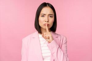 Hush, taboo concept. Portrait of asian businesswoman showing shush gesture, shhh sign, press finger to lips, standing over pink background in suit photo