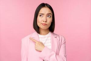 Doubtful business woman, sulking and grimacing while pointing, looking left at banner with skeptical face expression, posing against pink background photo