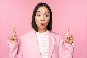 Portrait of asian businesswoman, corporate worker in suit, looks surprised by advertisement, points finger up, showing banner or logo on top, stands over pink background photo