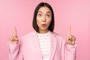 Enthusiastic corporate worker, asian business woman pointing fingers up and smiling, showing advertisement, logo, standing over pink background photo