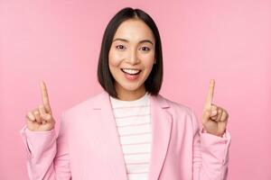 Smiling korean businesswoman, pointing fingers up, showing advertisement, banner or logo on top, standing in suit over pink background photo