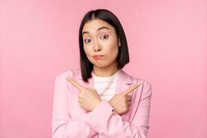 Decision concept. Young asian businesswoman, corporate worker pointing fingers sidways, two directions, showing variants, looking indecisive while choosing, pink background photo