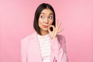 Portrait of asian corporate woman showing mouth seal, close shut lips on key gesture, promise keep secret, standing over pink background in suit photo