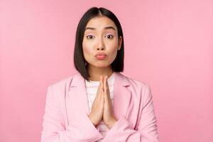 Portrait of asian businesswoman asking for help, say please, standing in praying, begging pose, pink studio background photo