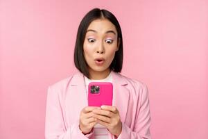 Portrait of asian businesswoman with surprised face, using smartphone app, wearing business suit. Korean girl with mobile phone and excited face expression, pink background photo