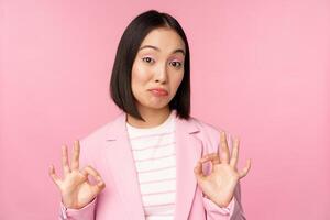 Not bad. Impressed businesswoman, asian office lady showing okay sign and nod in approval, standing over pink background photo