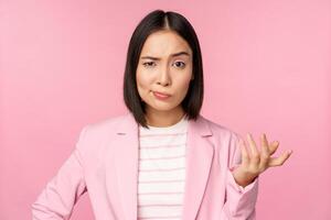 Portrait of angry asian woman in suit, clench fists and looking furious, outraged of smth bad, standing over pink background photo