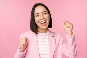 Enthusiastic saleswoman, asian corporate woman say yes, achieve goal and celebrating, triumphing, looking with rejoice and smiling, standing over pink background photo