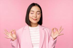 Keep calm. Smiling young asian woman meditating, practice yoga, mindfulness and relaxation at work, breath, inhale air with pleased face, standing over pink background photo