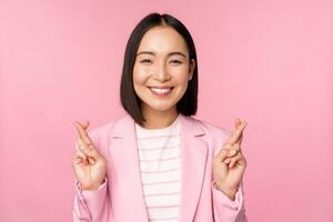 Happy asian businesswoman cross fingers for good luck, wishing, praying and hoping, smiling at camera, standing in suit over pink background photo