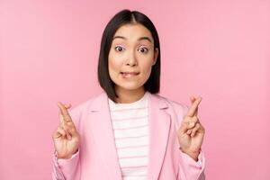 Happy lucky businesswoman, asian corporate lady wishing, making wish, hoping for smth and praying, standing in suit over pink background photo