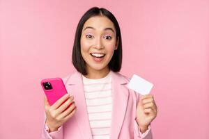 Image of smiling happy asian businesswoman showing credit card, paying online on smartphone application, order with mobile phon, standing against pink background photo