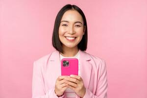 Portrait of smiling business woman, asian corporate person using smartphone, mobile phone application, standing over pink background photo