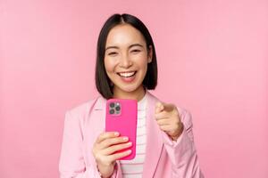 Happy asian businesswoman laughing, pointing finger at you and recording video, taking photos on smartphone, using mobile phone, pink background