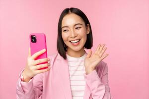 Stylish asian businesswoman, girl in suit taking selfie on smartphone, video chat with mobile phone app, posing against pink studio background photo