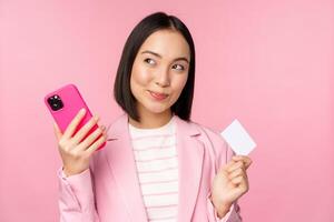 sonriente asiático corporativo mujer, dama en traje pensamiento, participación teléfono inteligente y crédito tarjeta, plan a comprar algo en línea, compras con móvil teléfono, rosado antecedentes foto