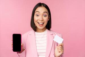 Enthusiastic asian businesswoman showing mobile phone screen and credit card, looking amazed at camera, standing over pink background photo