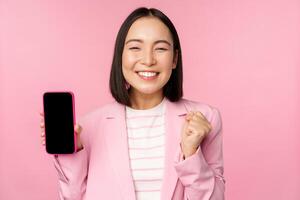 Winning korean businesswoman showing smartphone screen, smiling pleased, demonstrating mobile phone application, online store or shopping app, standing over pink background photo