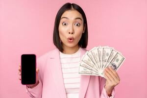 Image of korean successful corporate woman showing money, dollars and smartphone app screen, interface of mobile phone application, concept of investment and finance, pink background photo