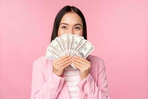 Happy asian lady in suit holding money, dollars with pleased face expression, standing over pink background photo