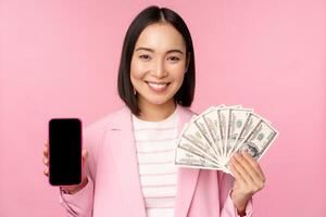 Image of korean successful corporate woman showing money, dollars and smartphone app screen, interface of mobile phone application, concept of investment and finance, pink background photo