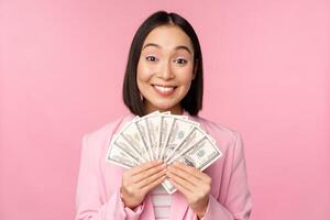 Finance, microcredit and people concept. Happy smiling asian businesswoman showing dollars money, standing in suit against pink background photo