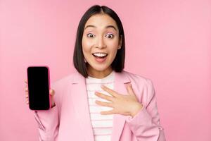 Surprised, enthusiastic asian businesswoman showing mobile phone screen, smartphone app interface, standing against pink background photo