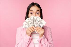 Happy asian lady in suit holding money, dollars with pleased face expression, standing over pink background photo