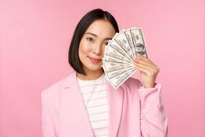 microcrédito, inversión y negocio personas concepto. joven asiático mujer de negocios, corporativo dama demostración dinero, efectivo dólares, sonriente complacido, rosado antecedentes foto