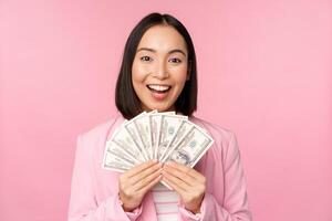 Finance, microcredit and people concept. Happy smiling asian businesswoman showing dollars money, standing in suit against pink background photo