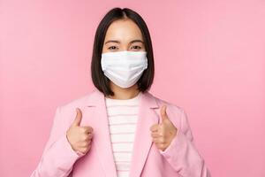 Asian businesswoman in suit and medical face mask, showing thumbs up, recommending wearing personal protective equipment in office during covid-19 pandemic, pink background photo