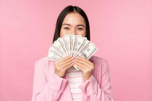 Happy asian lady in suit holding money, dollars with pleased face expression, standing over pink background photo