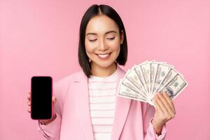 Image of korean successful corporate woman showing money, dollars and smartphone app screen, interface of mobile phone application, concept of investment and finance, pink background photo