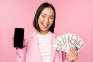 exitoso joven asiático mujer de negocios demostración dinero, efectivo dolares y teléfono inteligente pantalla, sonriente complacido, rosado antecedentes foto